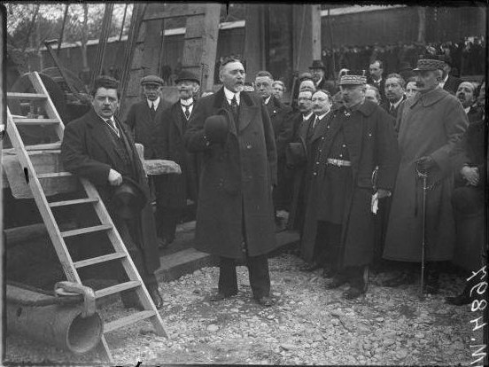Photographie ancienne en noir et blanc : Achille Lignon, debout au centre en manteau sombre, prononce un discours entouré d'officiels